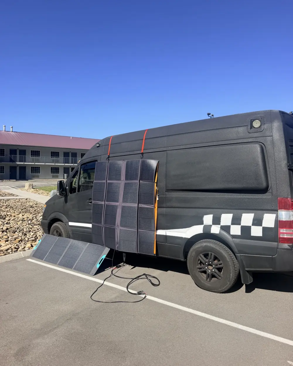 The black Rippin' van with folding solar panels hung on its side and laid out on the ground, charging under a clear blue sky.